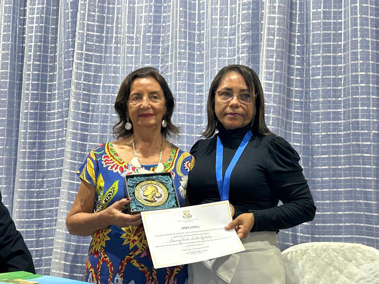 A profª Zenaide Santos Bayma, em nome de sua irmã Francy Anchieta, recebe a medalha e o diploma das mãos da Vereadora Ana Luiza Ramos, presidente da Câmara Municipal – Foto: Iago Azevedo