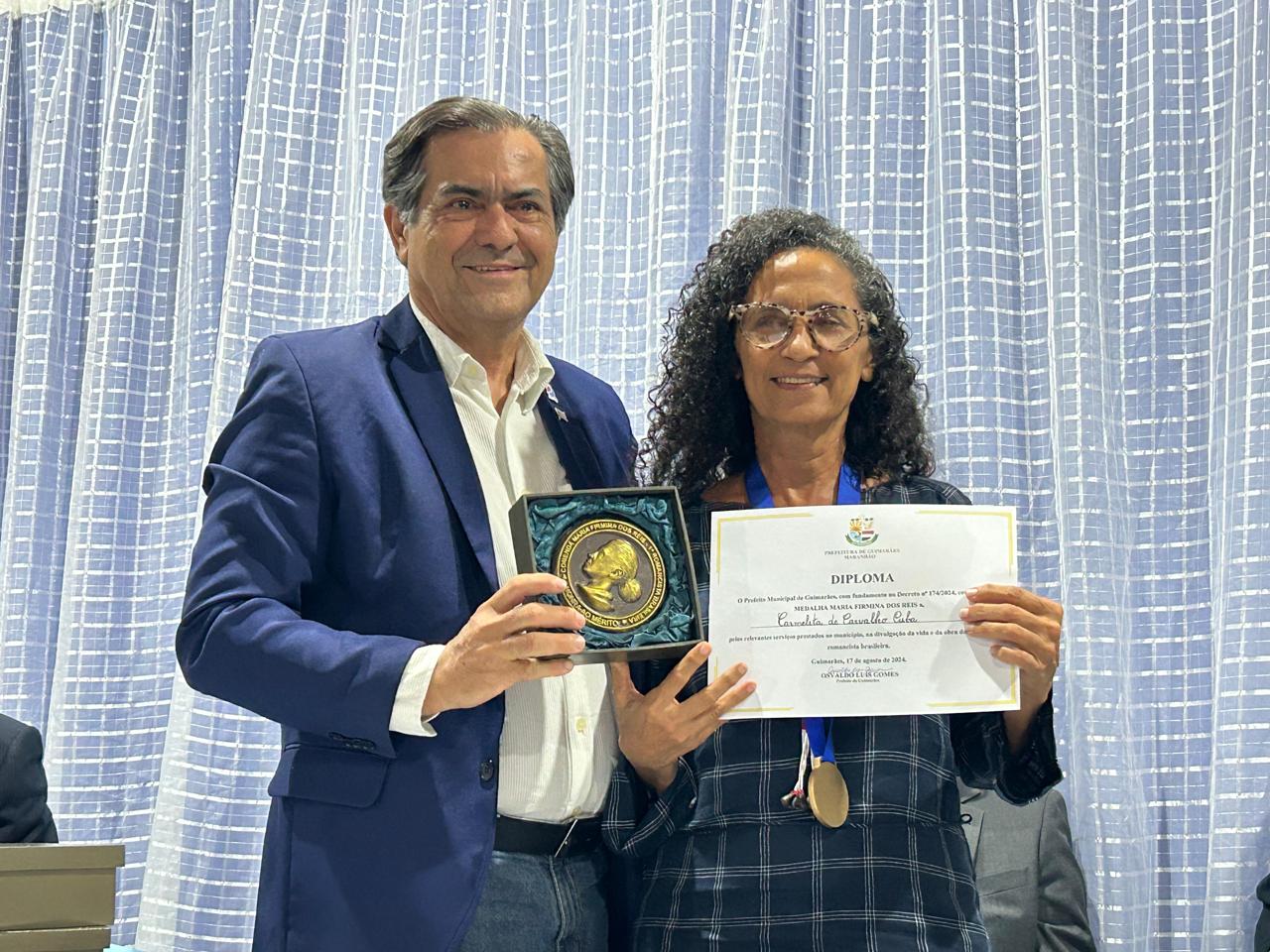 O escritor Luiz Tadheu Nunes e Silva entrega a medalha e o diploma à Profª Kátia Carvalho Cuba, que recebeu a honraria em nome de sua mãe Carmelita Carvalho Cuba, membro da Comissão do Sesquicentenário de nascimento de Maria Firmina dos Reis, em 1975 – Foto: Iago Azevedo